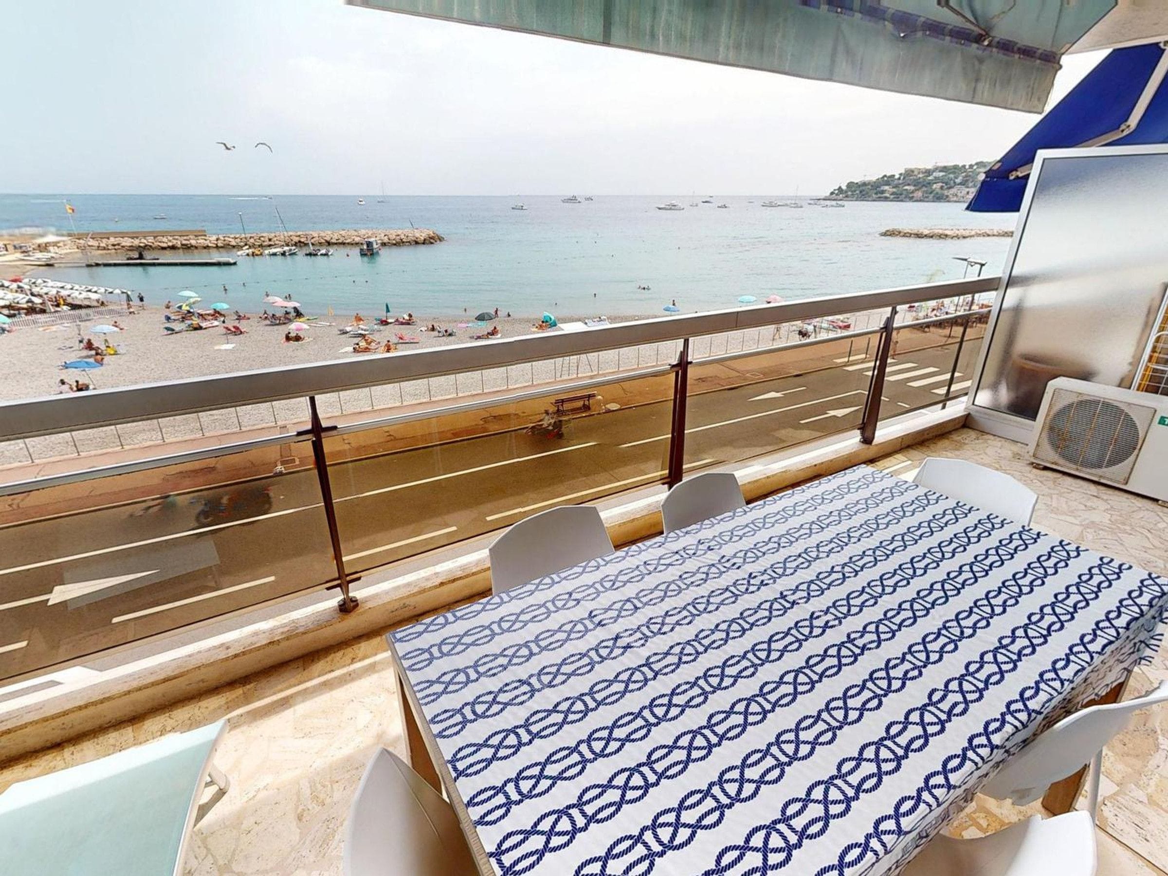 A balcony with a view of the beach and sea, a table with a patterned tablecloth, and a row of chairs.