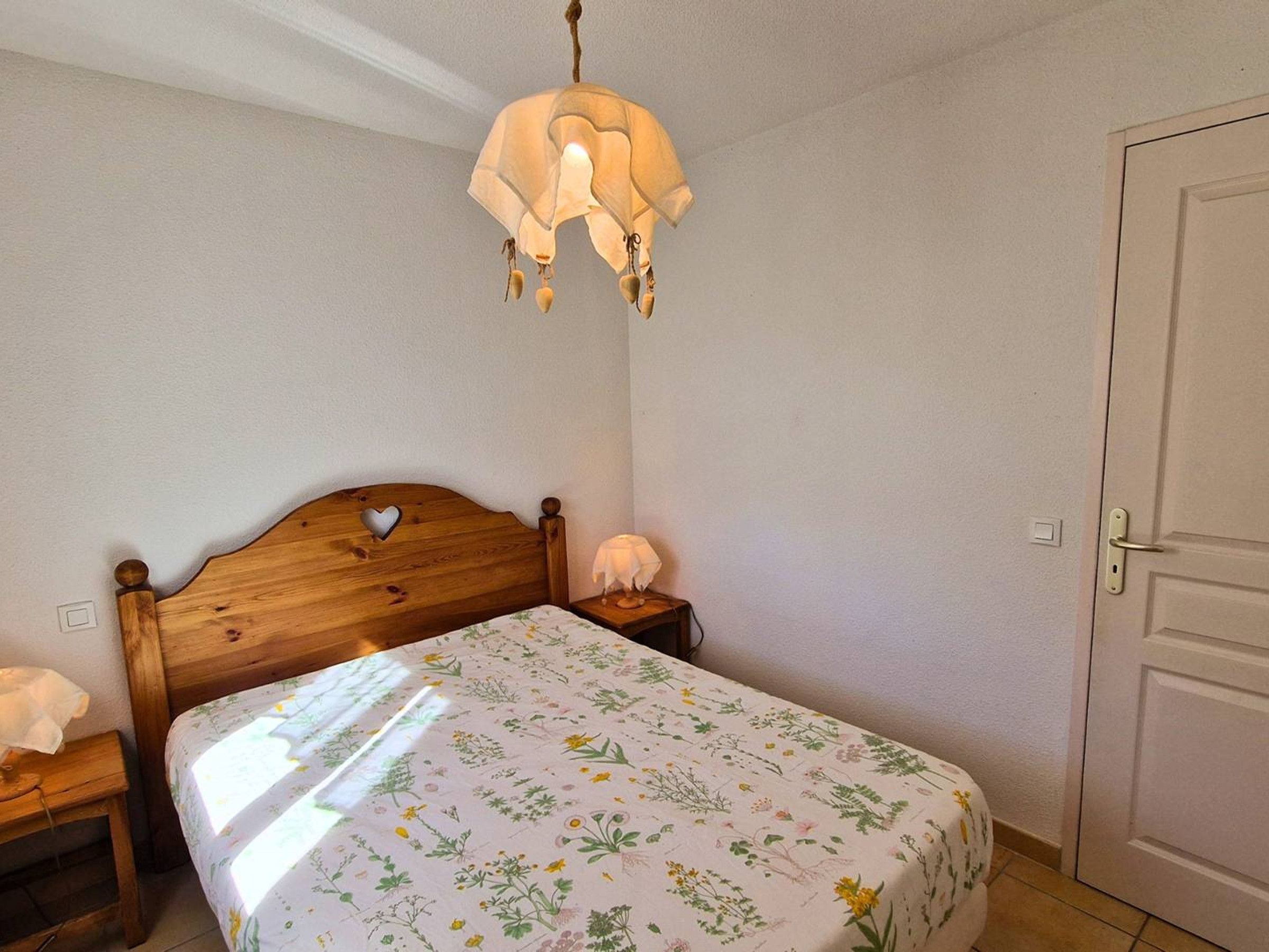 Een slaapkamer met een houten bed, een nachtkastje, een wandlamp en een deur.