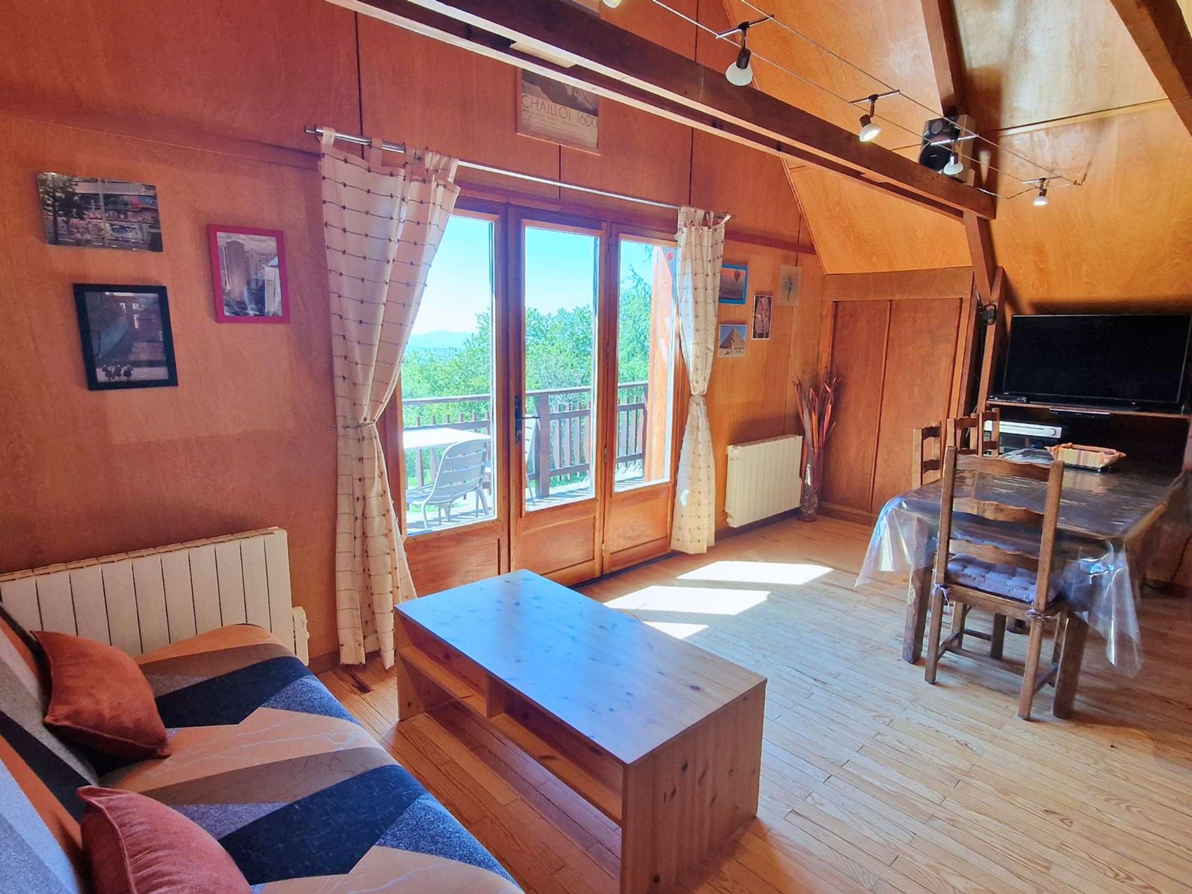 Interior acogedor de una cabaña con una mesa de comedor de madera, un sofá con cojines estampados, un televisor y un amplio ventanal con cortinas.