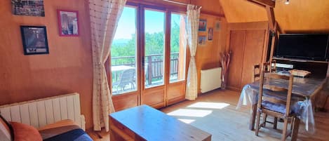 Ein gemütliches Blockhaus-Interieur mit einem Holzesstisch, einer Couch mit gemusterten Kissen, einem Fernseher und einem großen Fenster mit Vorhängen.