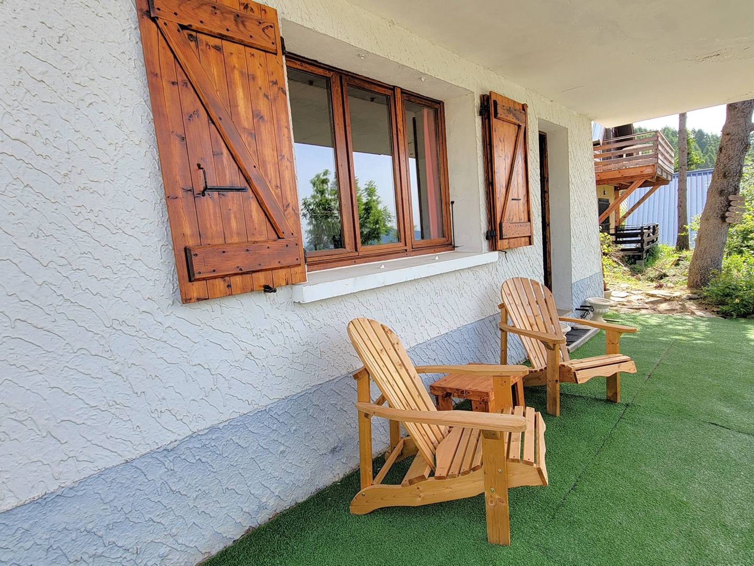 Een gebouw met houten luiken, een raam en twee houten stoelen op een groene ondergrond.
