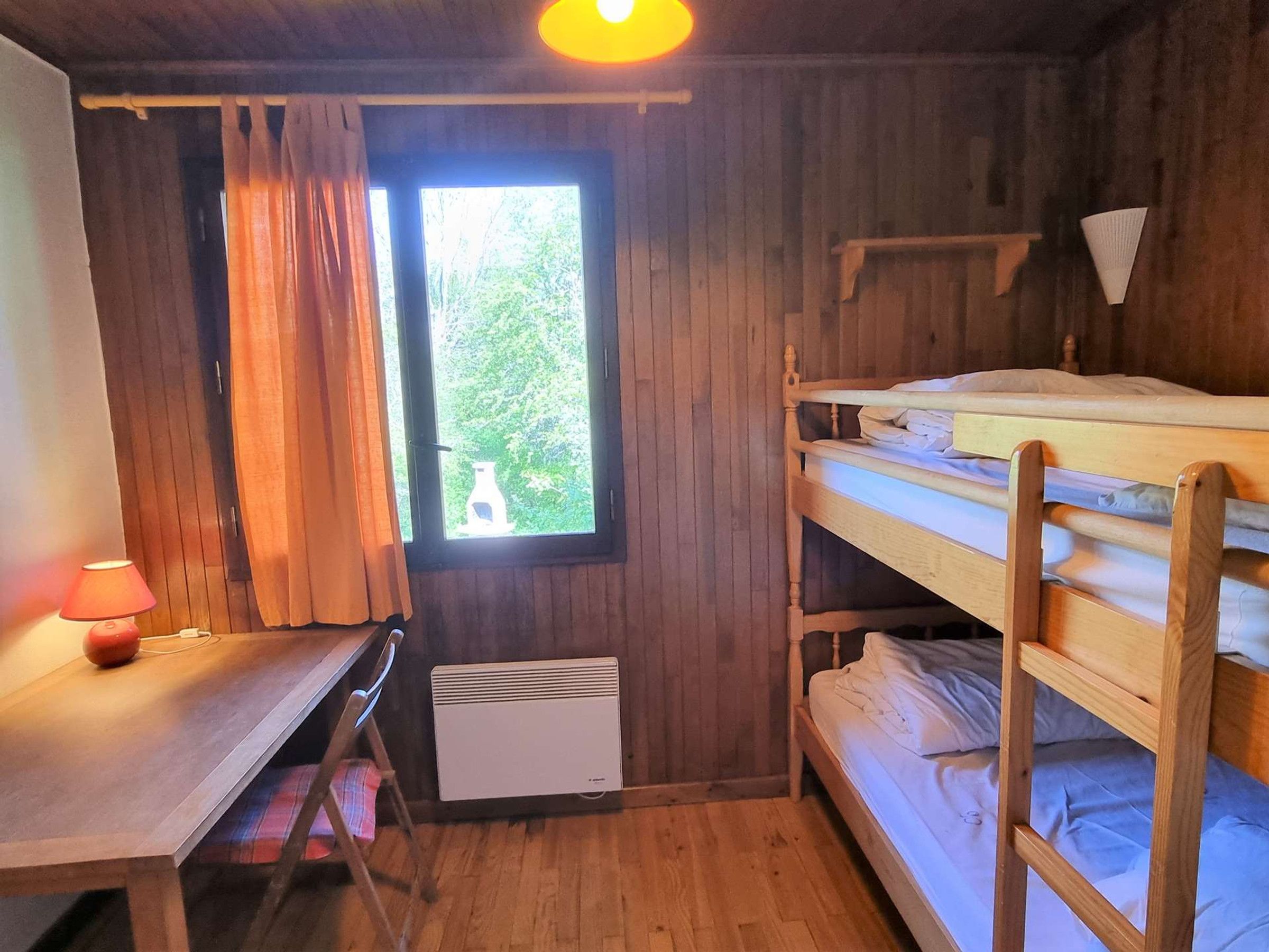 A room with a bunk bed, a desk with a lamp, a window with curtains, and a radiator.