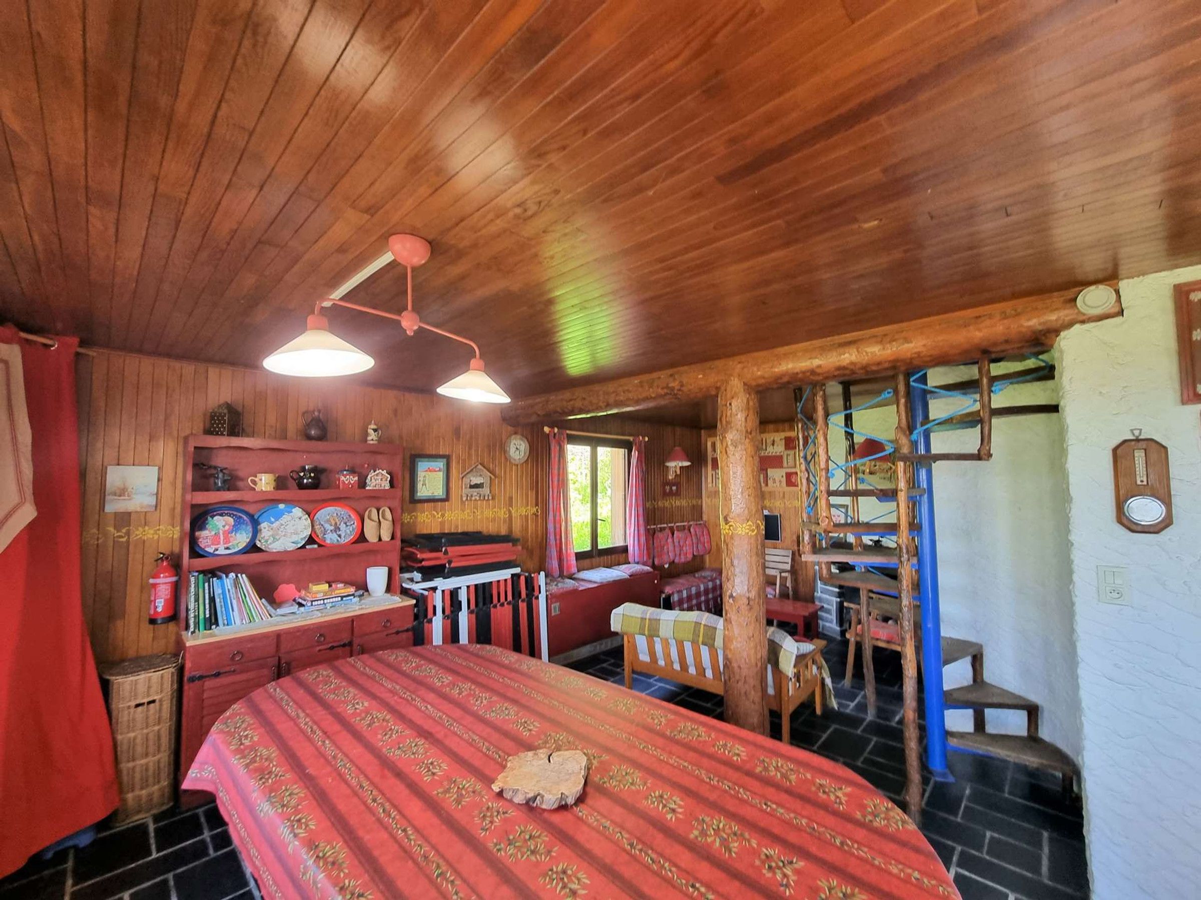 A cozy cabin interior with a wooden ceiling, a bed with a red patterned bedspread, a small desk, and a chair.