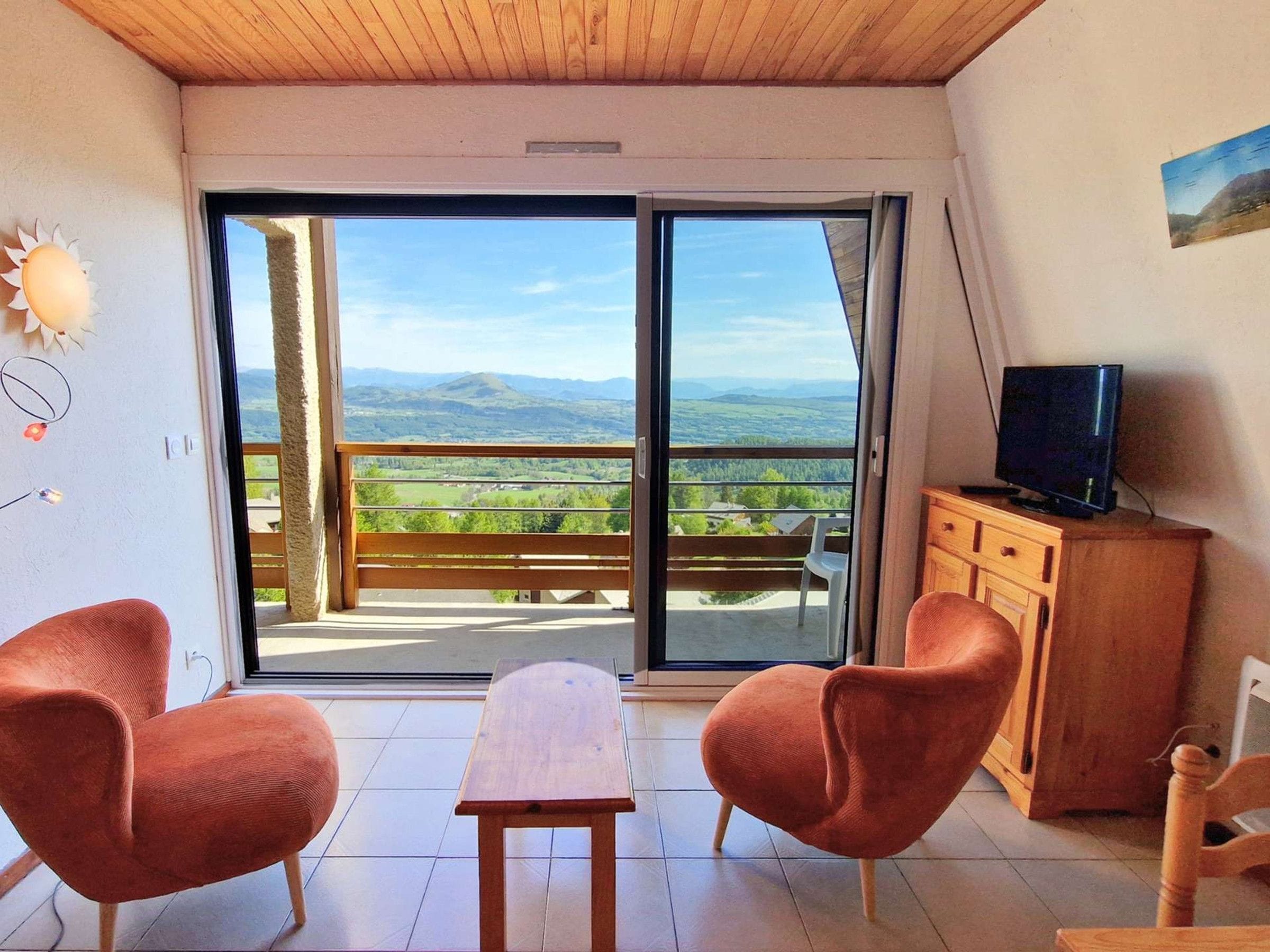 A room with a wooden ceiling, a large sliding glass door leading to a balcony with a view of a mountainous landscape, two orange chairs, a small wooden table, and a television on a wooden cabinet.