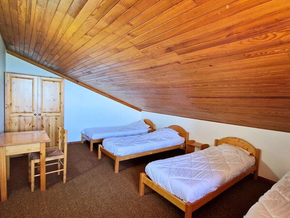 A small, single-room accommodation with a sloped ceiling, two wooden beds, a wooden desk, and a chair.