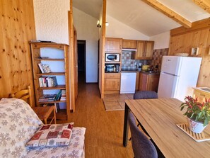 A cozy room with a wooden bookshelf, a dining table, and a kitchen area with wooden cabinets and appliances.