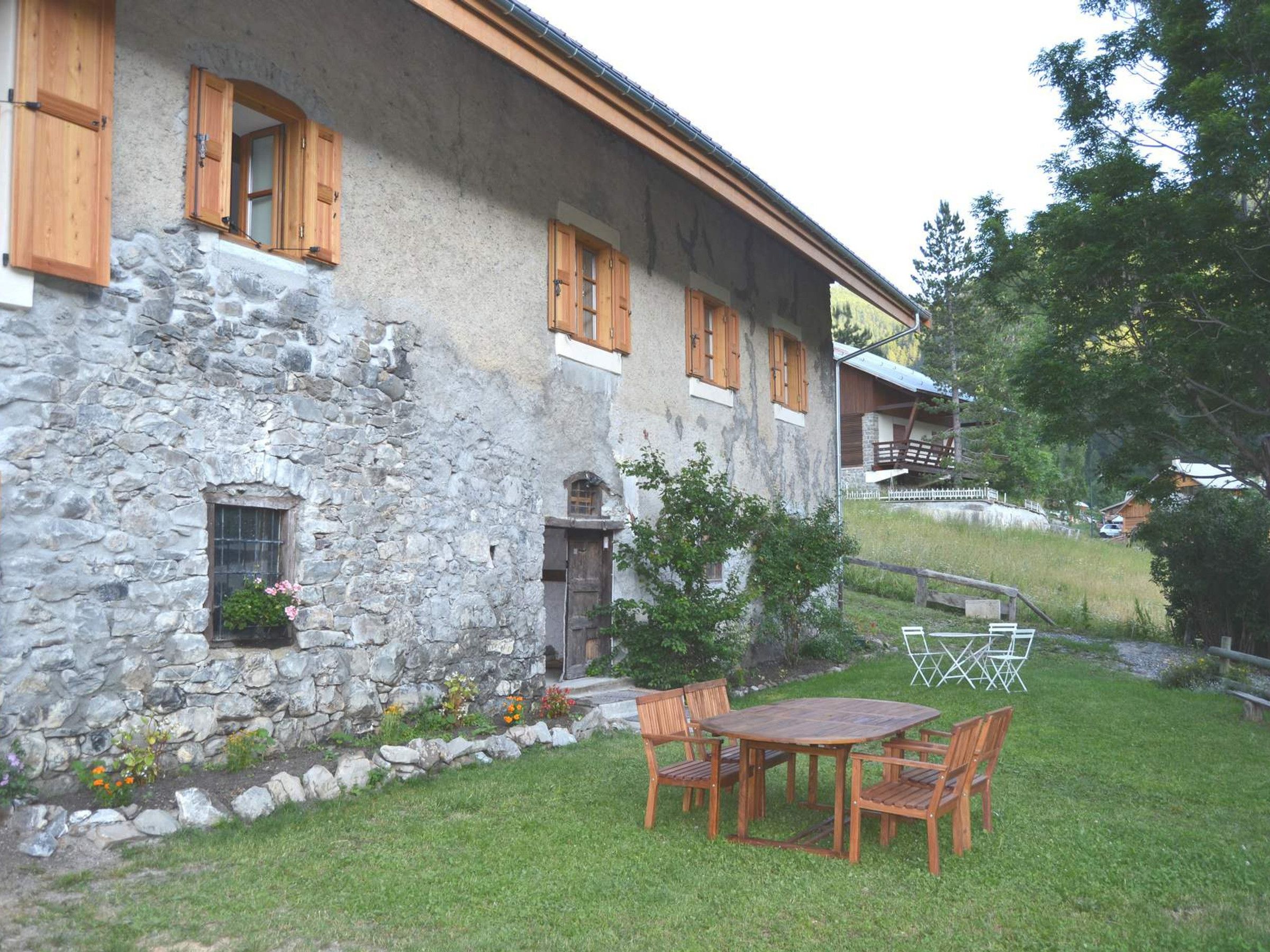 A stone building with wooden shutters, a small garden with a table and chairs, and a wooden deck.