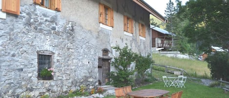 A stone building with wooden shutters, a small garden with a table and chairs, and a wooden deck.