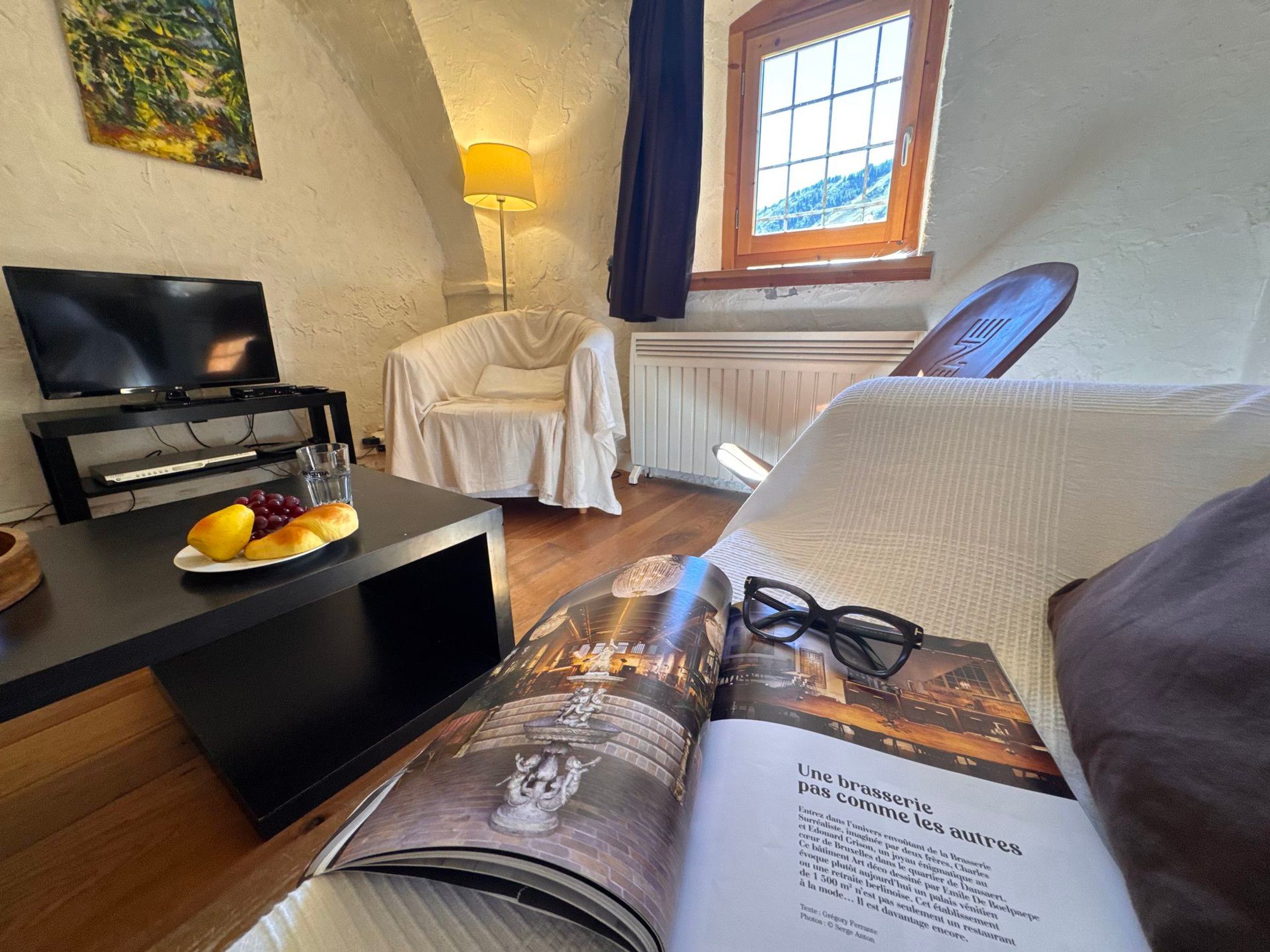 Une pièce avec une télévision, une table garnie de fruits, un magazine, une chaise blanche et une fenêtre donnant sur un beau paysage.