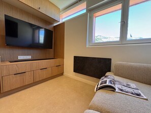 A modern living room with a flat-screen TV mounted on a wooden cabinet, a sofa with a magazine, and a window with a view of a landscape. - Apartment in the heart of Vars - 4 guests (Vars)