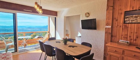 Zona pranzo con un tavolo grande e sedie, una televisione montata a parete e vista sul paesaggio attraverso ampie finestre.