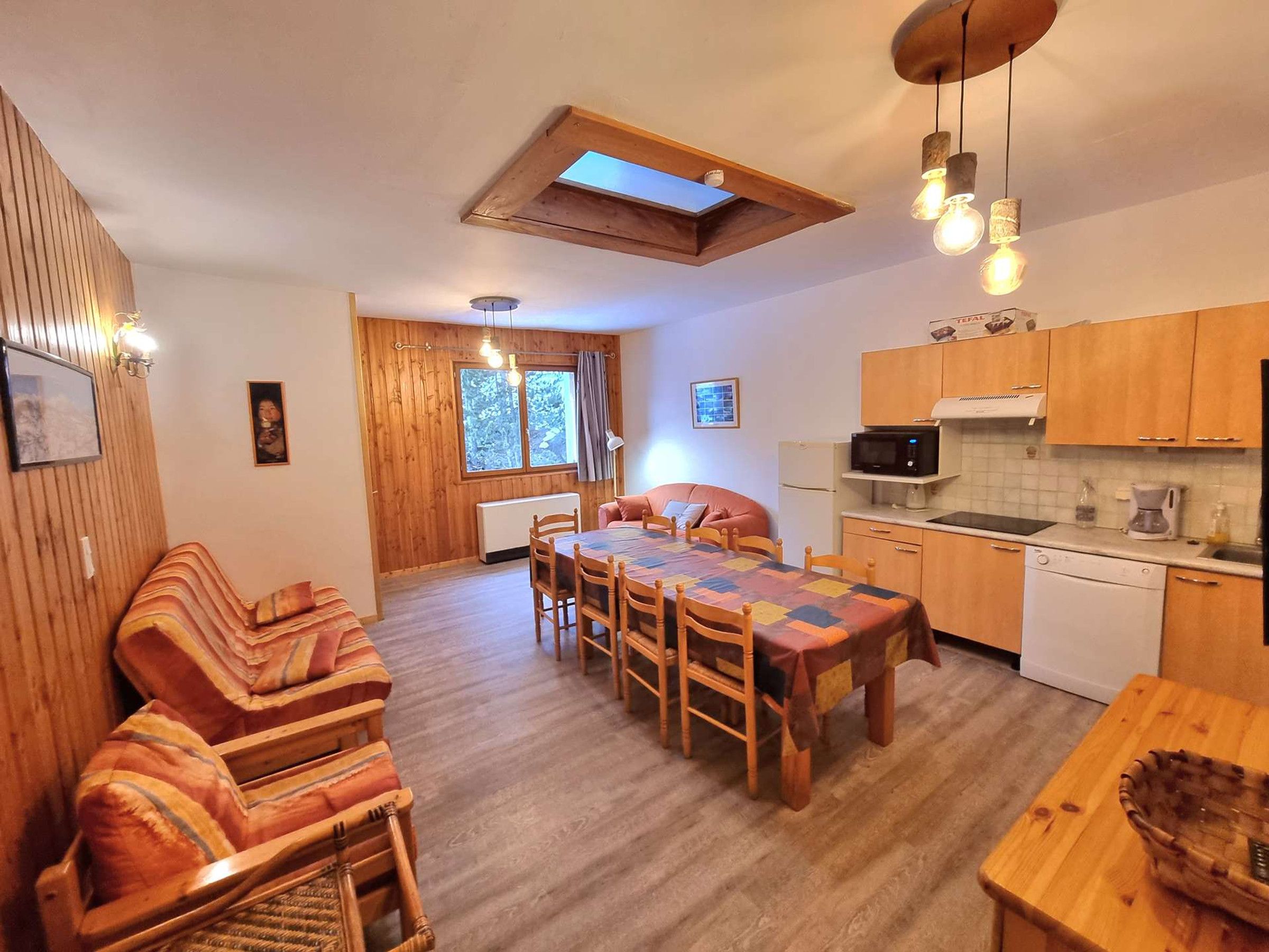 A cozy room with a wooden dining table, chairs, a sofa, a kitchen area with cabinets and appliances, and a skylight above the table.