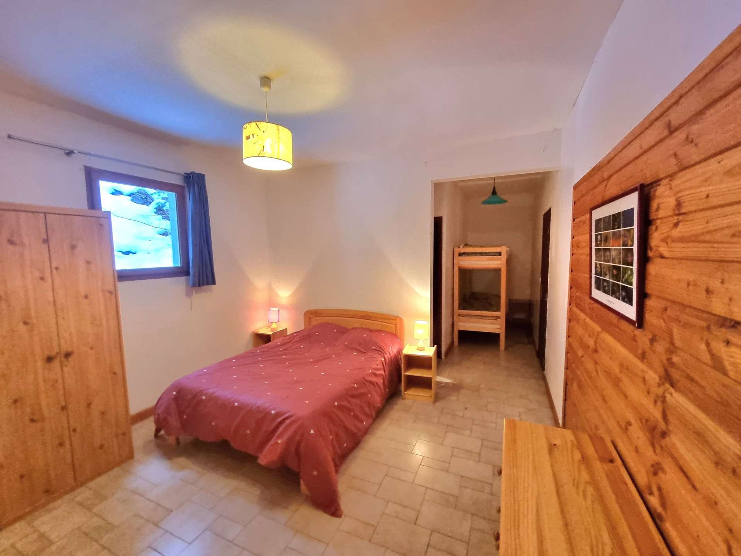 Een slaapkamer met een bed, nachtlampjes, een houten garderobe, een raam met gordijnen en een houten wand met een lijstwerk.