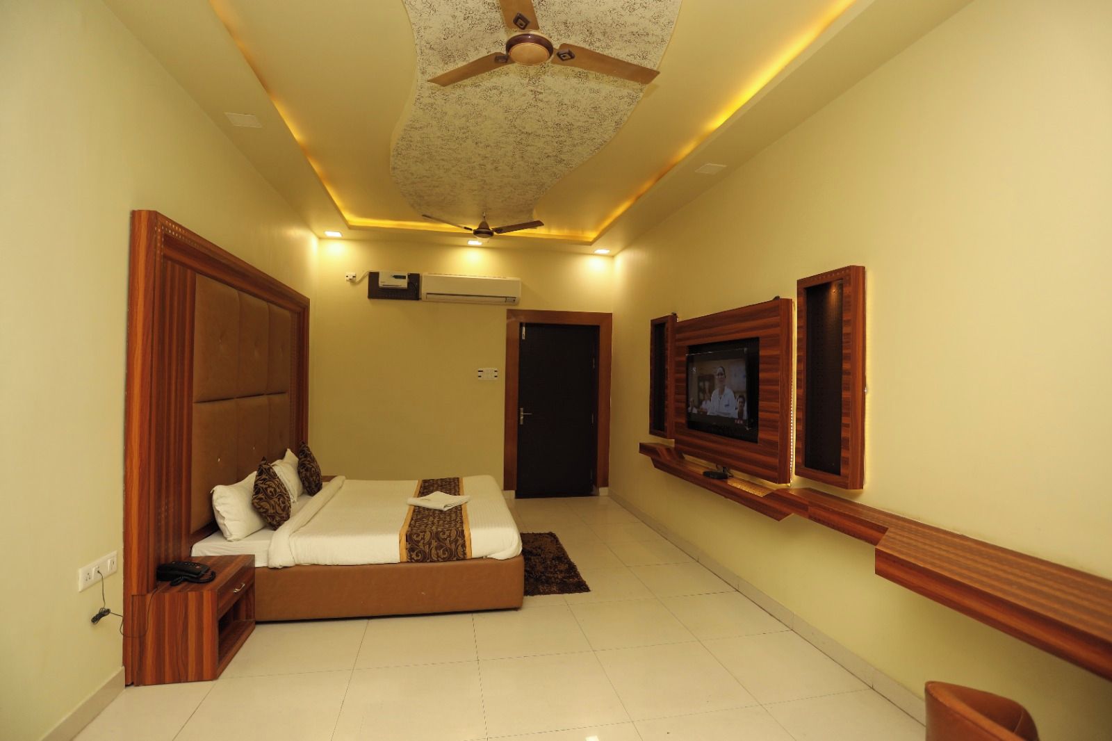 A hotel room with a bed, a television mounted on the wall, a ceiling fan, and a door.