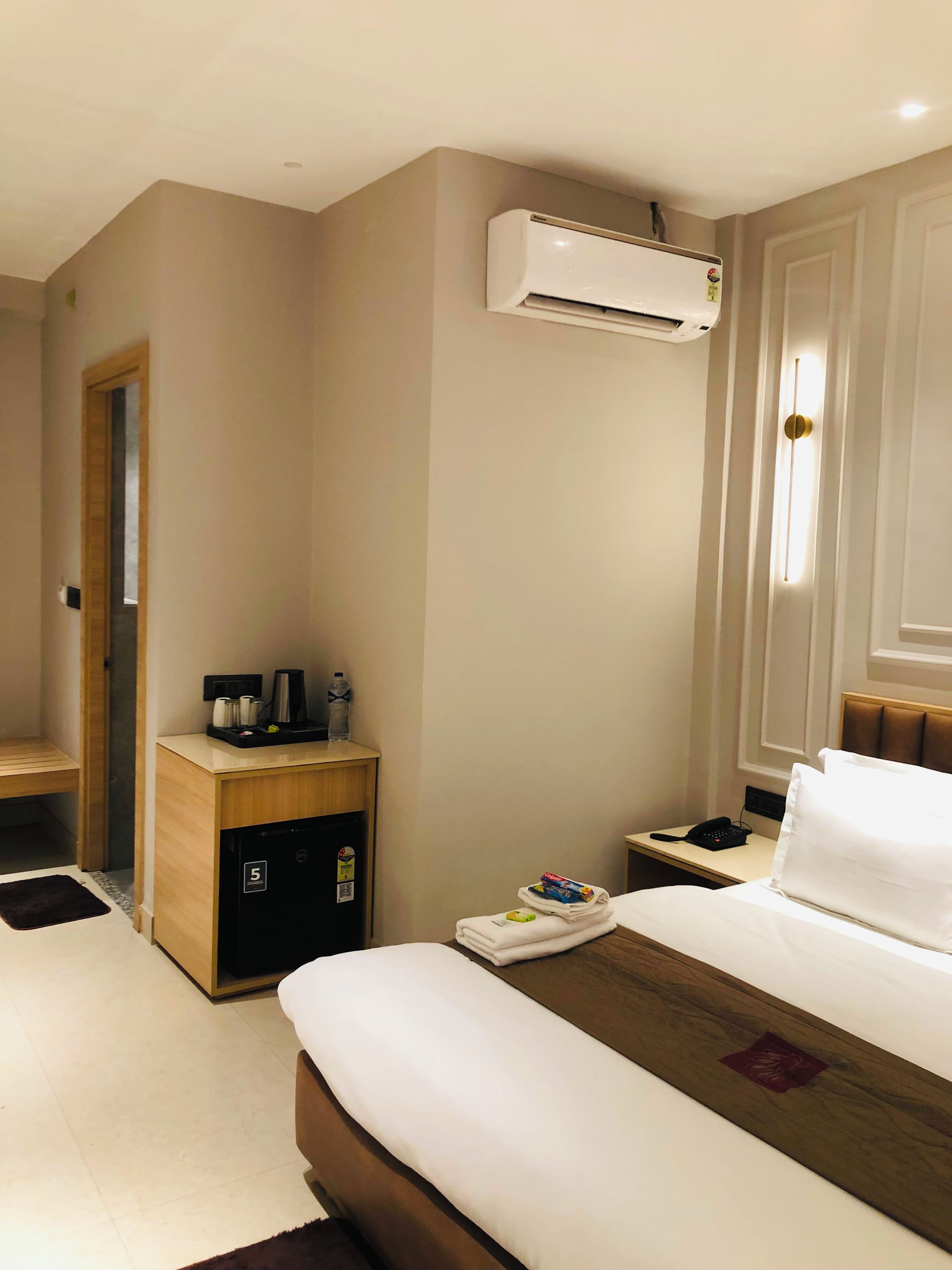 A hotel room with a bed, a bedside table, a small cabinet, an air conditioning unit, a nightstand, and a wall-mounted light.