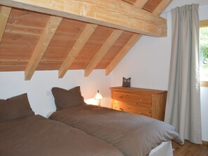 Ein Schlafzimmer mit schrägem Dach, Holzbalken, einem Schminktisch, einer Lampe und einem Fenster mit Vorhängen.
