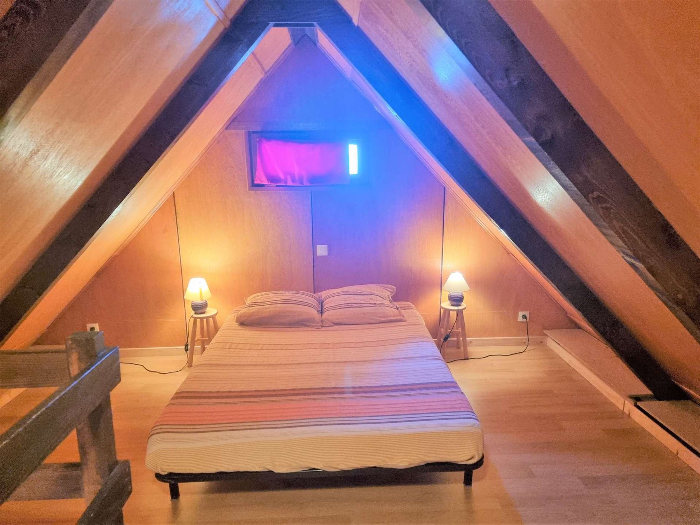 Ein gemütliches Schlafzimmer im Dachgeschoss mit schrägen Wänden, einem Bett mit gestreifter Bettdecke, zwei Nachttischlampen und einem angebrachten Fernseher.