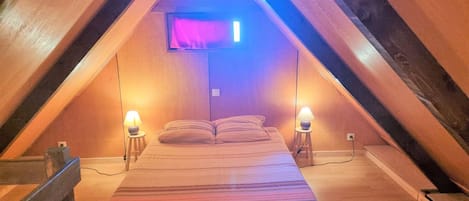 Ein gemütliches Schlafzimmer im Dachgeschoss mit schrägen Wänden, einem Bett mit gestreifter Bettdecke, zwei Nachttischlampen und einem angebrachten Fernseher.