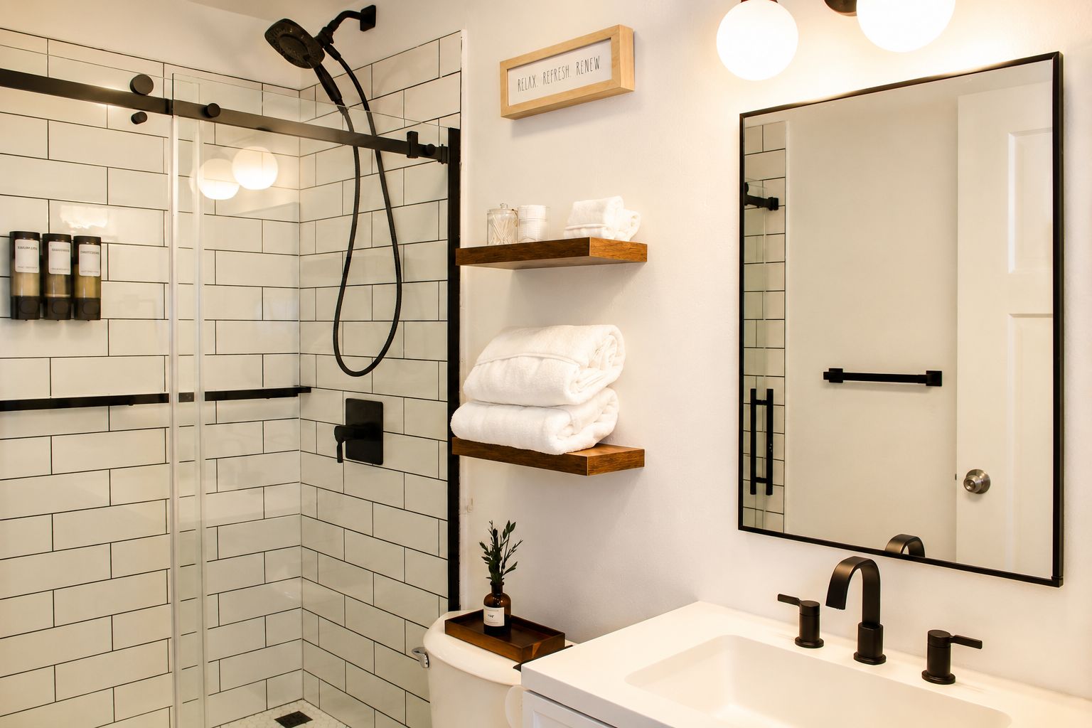 A modern bathroom with a glass shower enclosure, a white sink with black faucets, and a mirror with a wooden shelf above it.