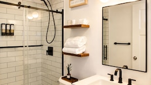 Une salle de bain moderne avec une douche à l’italienne en verre, un lavabo blanc doté de robinets noirs et un miroir surmonté d’une étagère en bois.