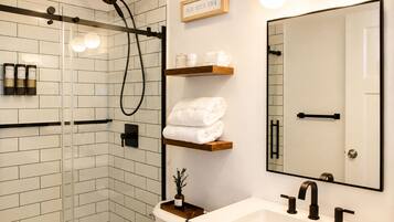 A modern bathroom with a glass shower enclosure, a white sink with black faucets, and a mirror with a wooden shelf above it.
