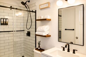 A modern bathroom with a glass shower enclosure, a white sink with black faucets, and a mirror with a wooden shelf above it.
