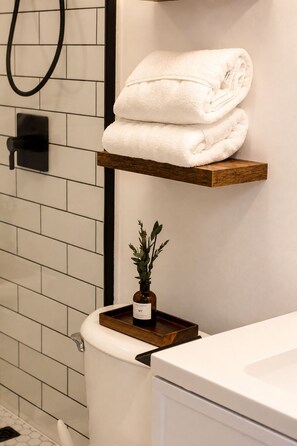 A bathroom with a toilet, a shower, and a sink. There is a towel rack with folded towels and a small plant on a tray.