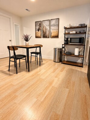 A dining area with a wooden table and chairs, a trash can, a microwave, and a shelf with kitchenware.