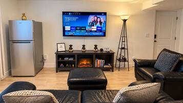 Een moderne woonkamer met een flatscreen televisie aan de muur, een donker houten dressoir eronder, een zwarte leren bank, een klein tafeltje met decoratieve objecten, en een vloerlamp.