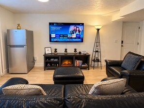 A modern living room with a flat-screen TV mounted on the wall, a dark wooden entertainment unit below, a black leather sofa, a small table with decorative items, and a standing floor lamp.