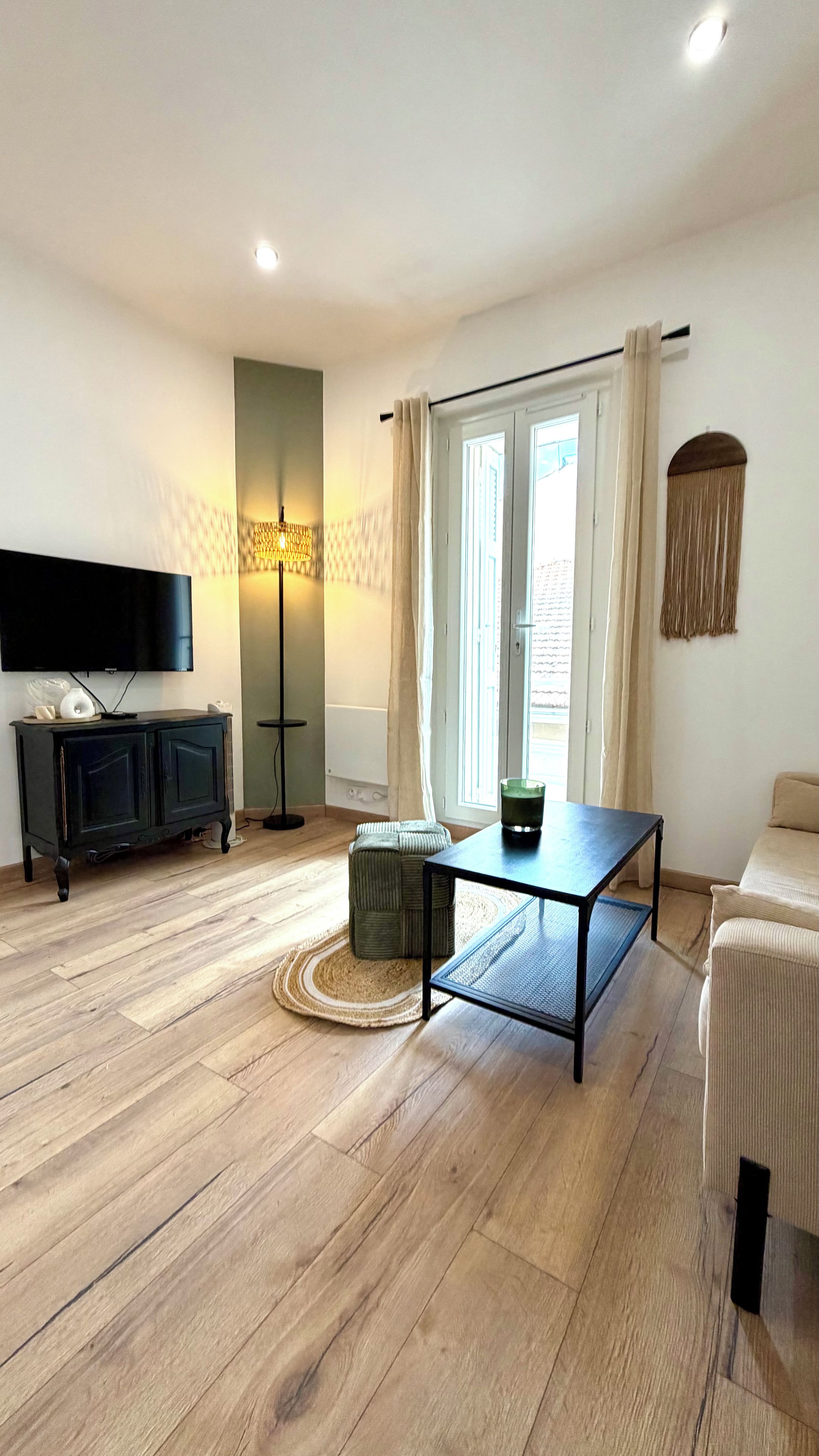 Un salon équipé d’un téléviseur à écran plat, d’un meuble TV noir, d’une table basse en verre, d’un canapé beige et d’une grande fenêtre avec des rideaux.