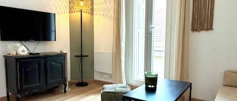 Una sala de estar con televisor de pantalla plana, un mueble de TV negro, una mesa de centro de vidrio, un sofá color beige y un ventanal con cortinas.