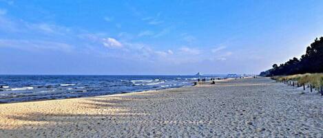 A wide sandy beach with gentle waves, a clear sky, and a distant pier.