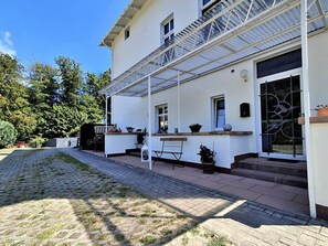 Ein weißes Gebäude mit überdachter Terrasse, Sitzgelegenheiten im Freien und einem gepflasterten Weg.