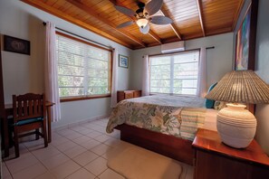 Ein Schlafzimmer mit Bett, Schreibtisch, Stuhl, Deckenventilator und zwei Fenstern mit Jalousien.
