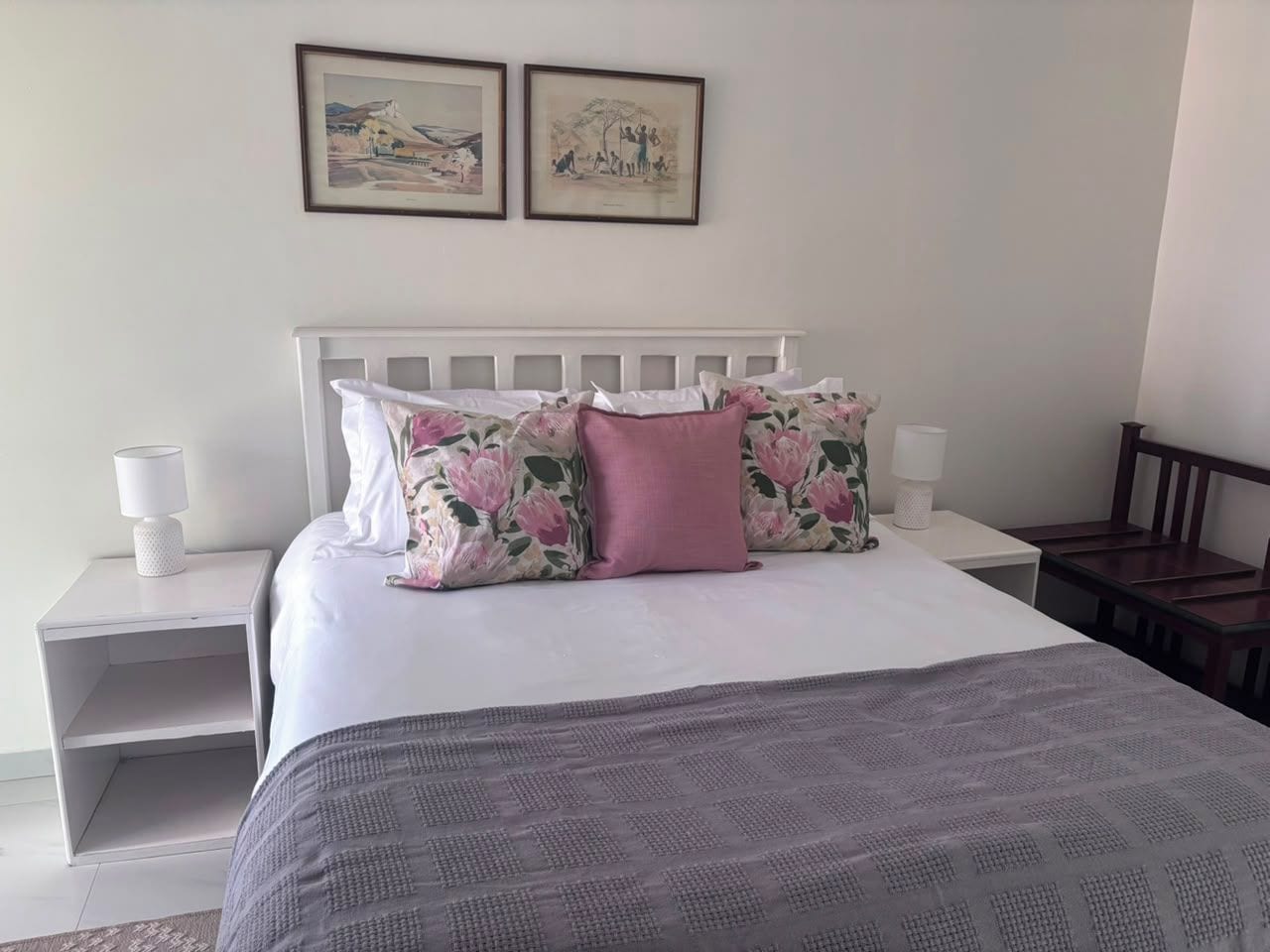 Un dormitorio con cama, mesitas de noche blancas, cabecera y dos cuadros enmarcados en la pared.