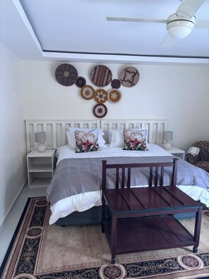 Ein Schlafzimmer mit einem Bett, zwei Nachttischen, einem Holzstuhl, einem Deckenventilator und dekorativen Tellern an der Wand.