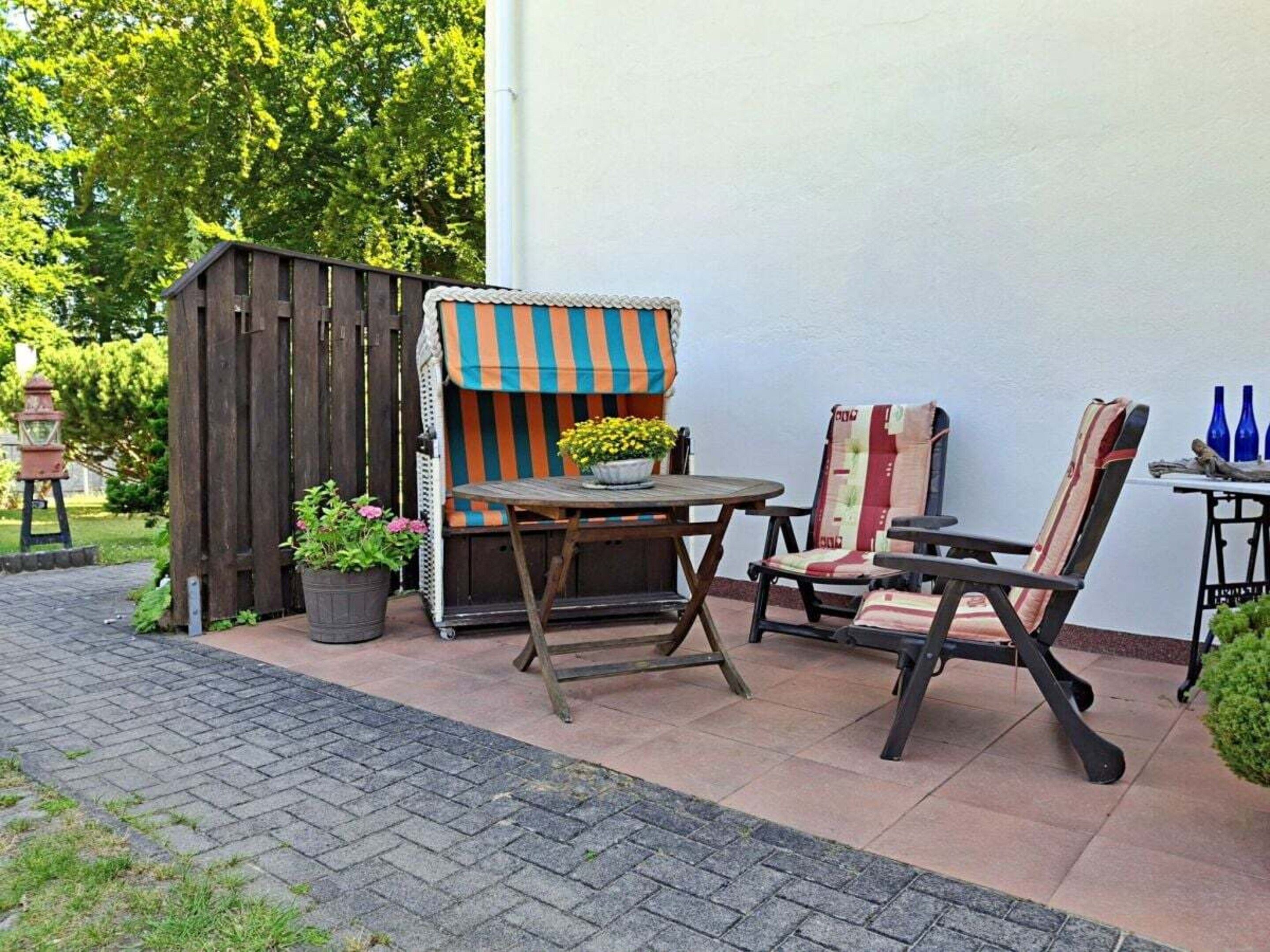 Een terras met een houten tafel en twee stoelen, een klein houten tuinhuisje, en een bloempot met een plant.