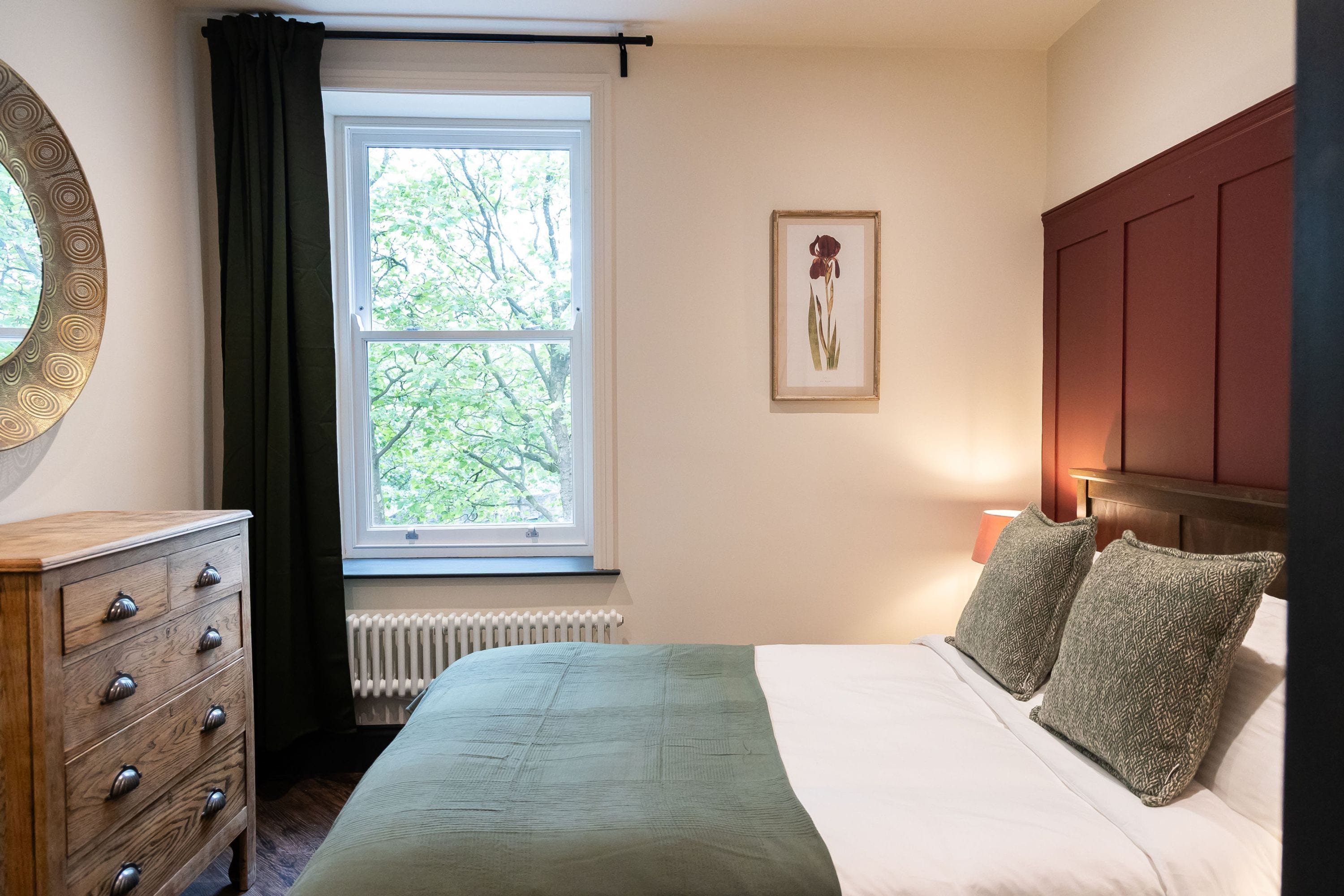 A bedroom with a bed, a dresser, a window with curtains, a radiator, and a framed picture on the wall.
