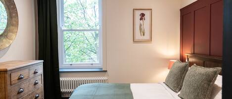 A bedroom with a bed, a dresser, a window with curtains, a radiator, and a framed picture on the wall.
