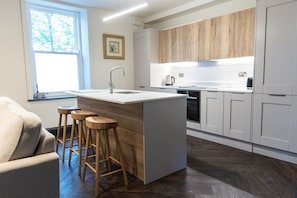 A modern kitchen with a central island, wooden stools, and a window with a view of trees. - Summerhill in Newcastle (Newcastle-upon-Tyne)
