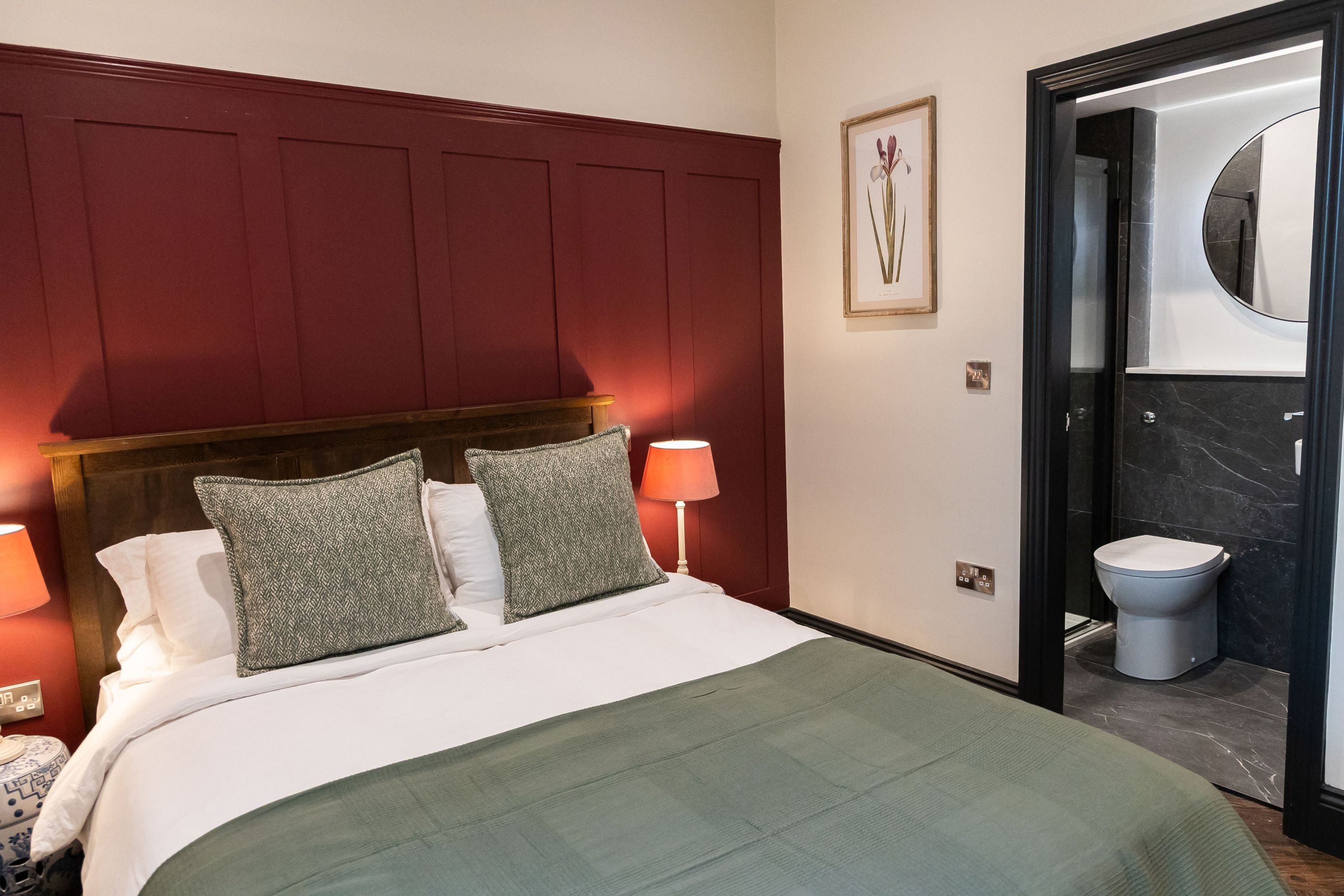 Habitación de hotel con cama, dos lámparas de noche, un cuadro en la pared y un baño visible a través de una puerta abierta.