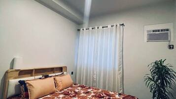 A bedroom with a bed, a bedside table, a plant, and a window with curtains.