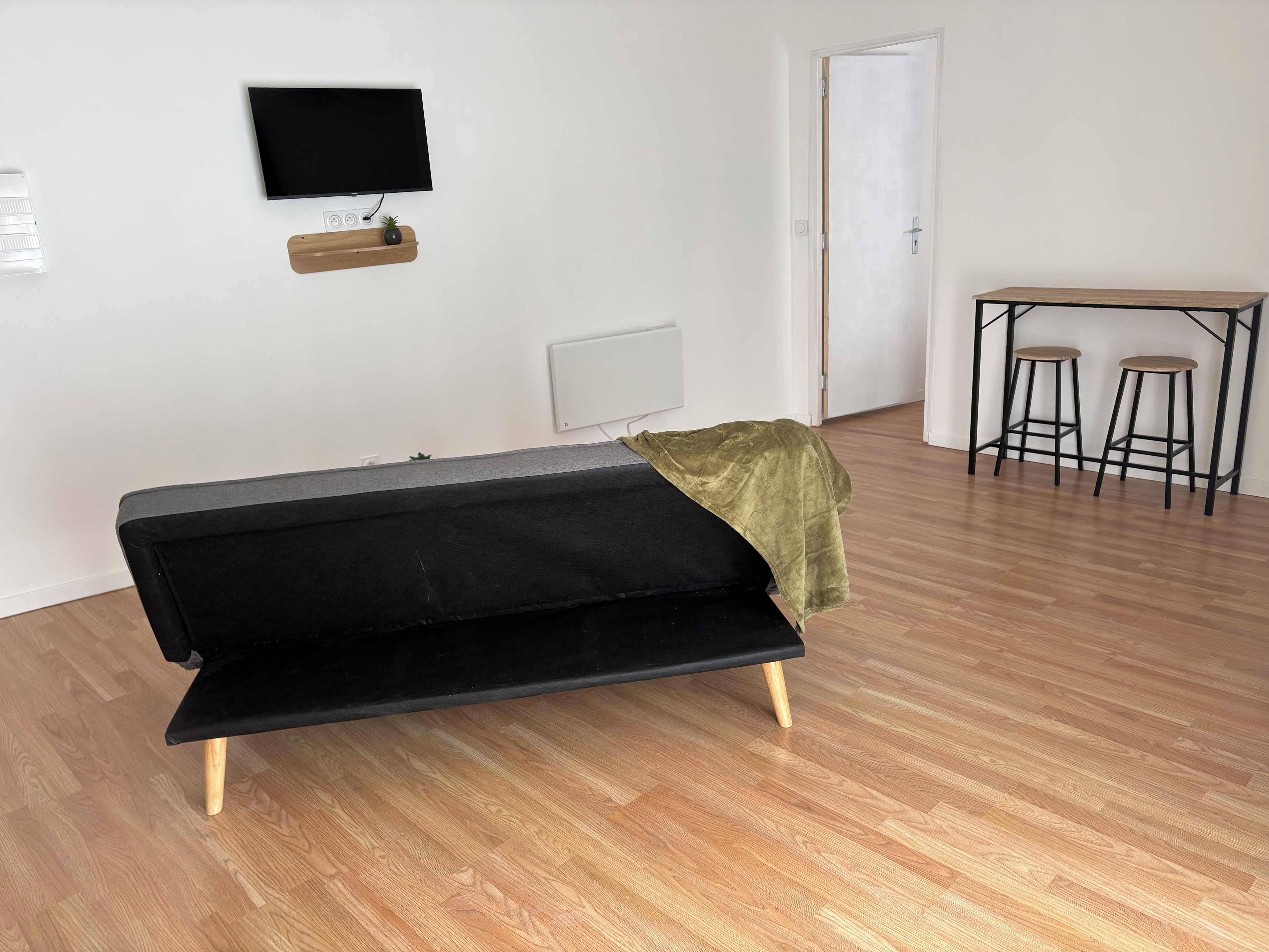 Une pièce avec un canapé noir, une petite table accompagnée de deux tabourets, un téléviseur fixé au mur et une porte.