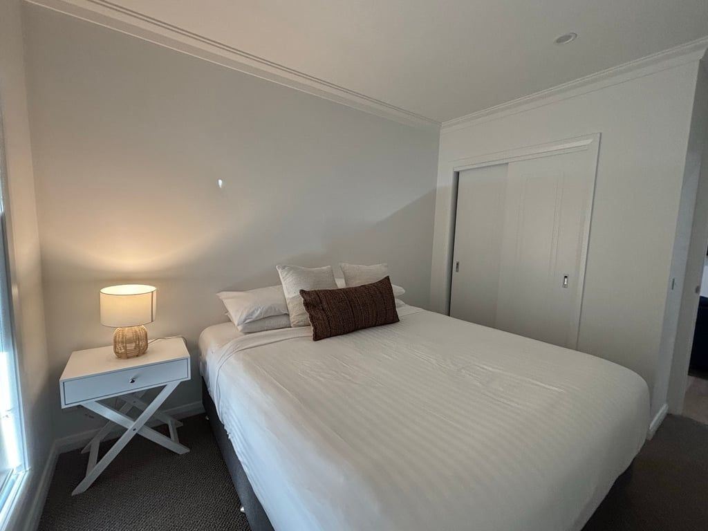 A hotel room with a single bed, a nightstand with a lamp, a door, and a window.