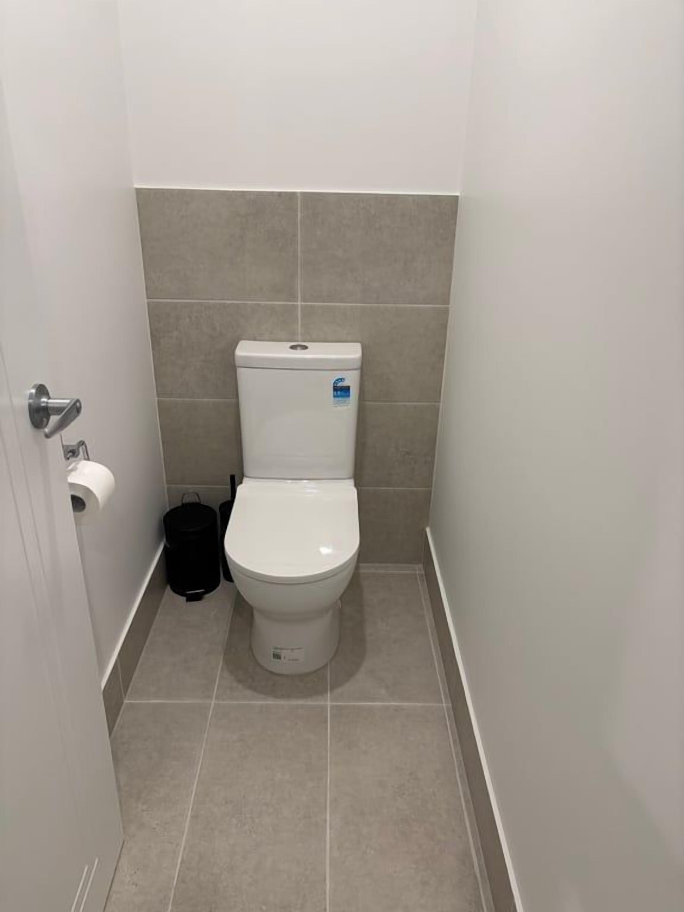 A small, narrow bathroom with a white toilet, a black trash bin, and a roll of toilet paper on the wall.