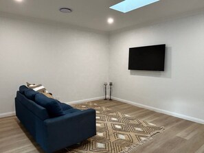 Quarto com sofá azul escuro, tapete estampado, televisão na parede e clarabóia.
