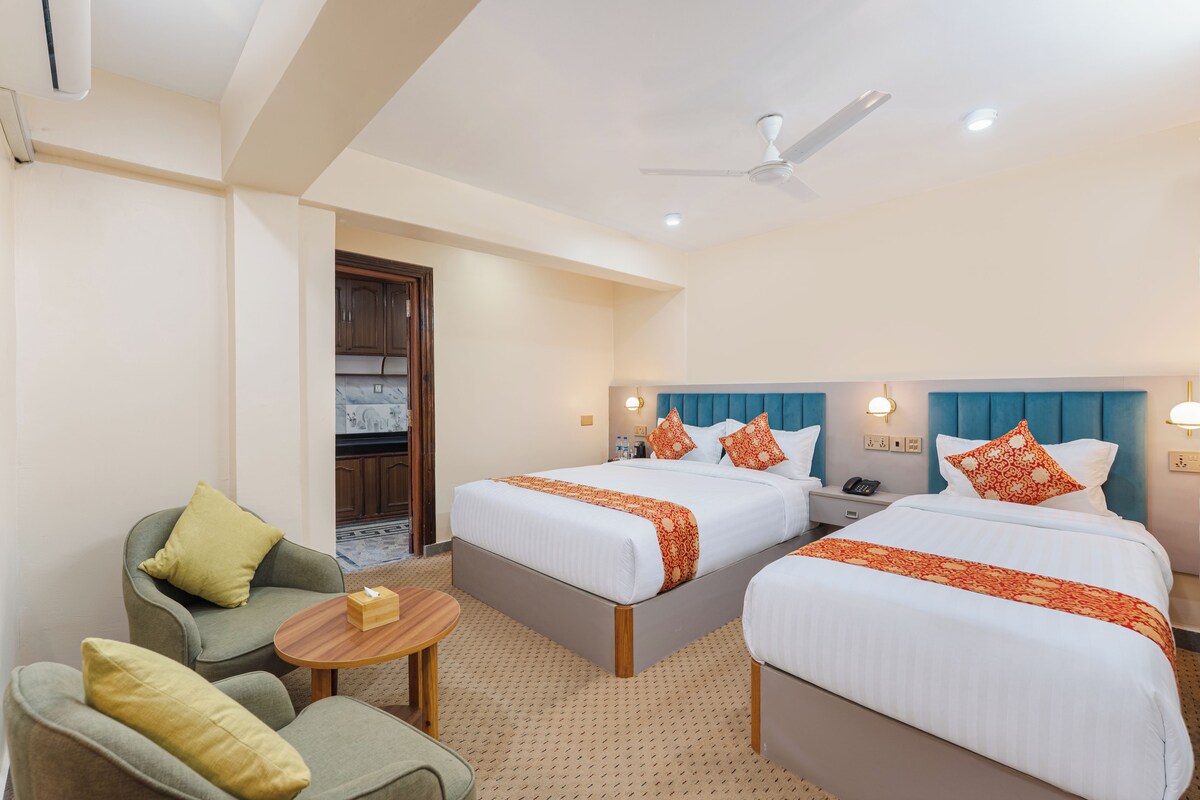 Habitación de hotel con dos camas, una zona de estar con una silla y una mesa, ventilador de techo y kitchenette.