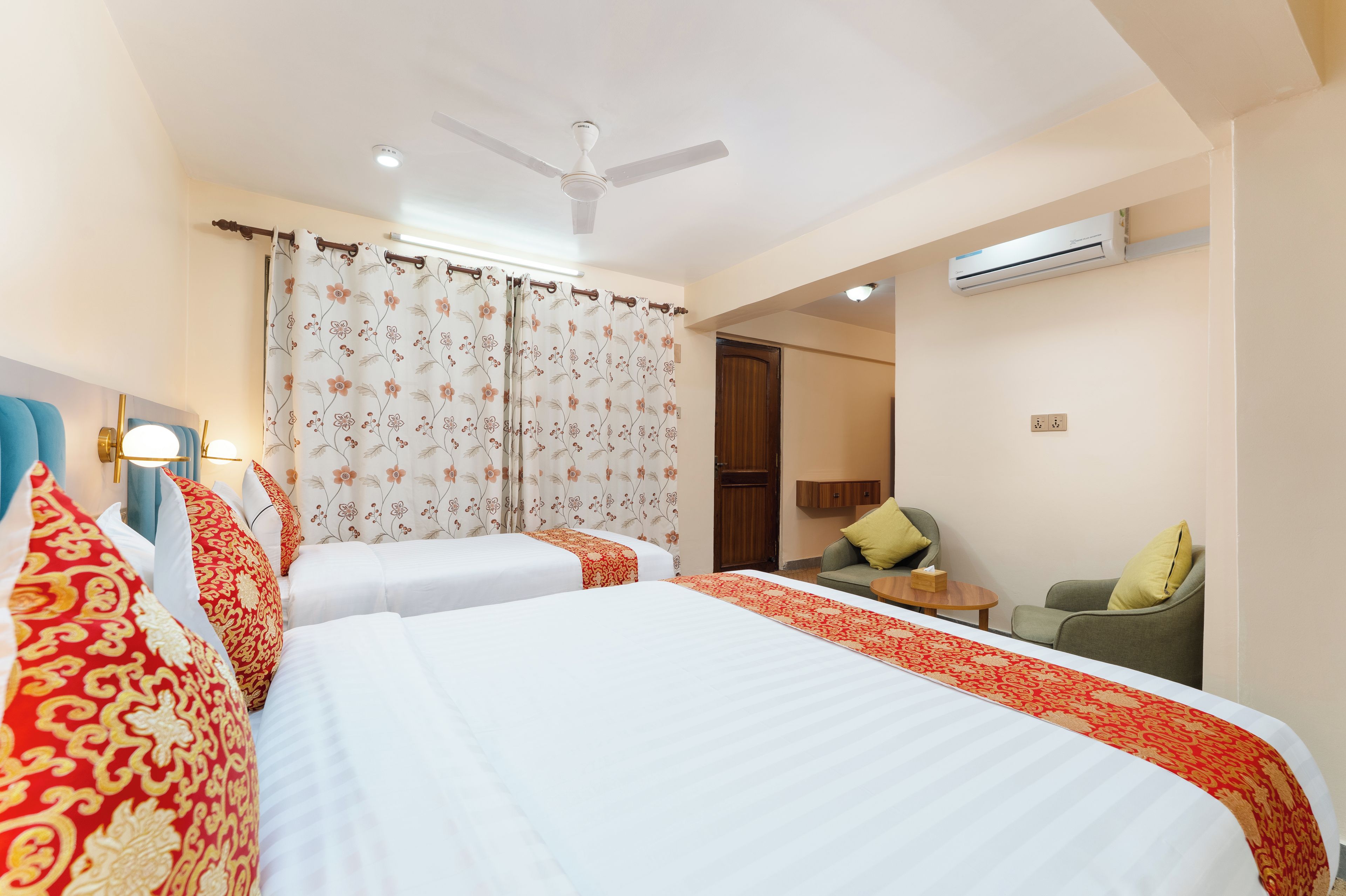 Habitación de hotel con dos camas, ventilador de techo, aire acondicionado, una pequeña mesa y un rincón de estar con sofá y sillones.