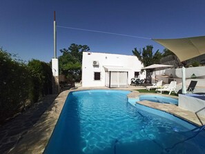 Una piscina amb una tenda de sol i gandules, envoltada d'un edifici de color blanc i vegetació.